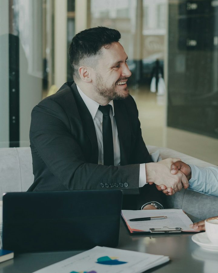 A salesman in a suit shaking hands with a customer across the desk of a car dealership