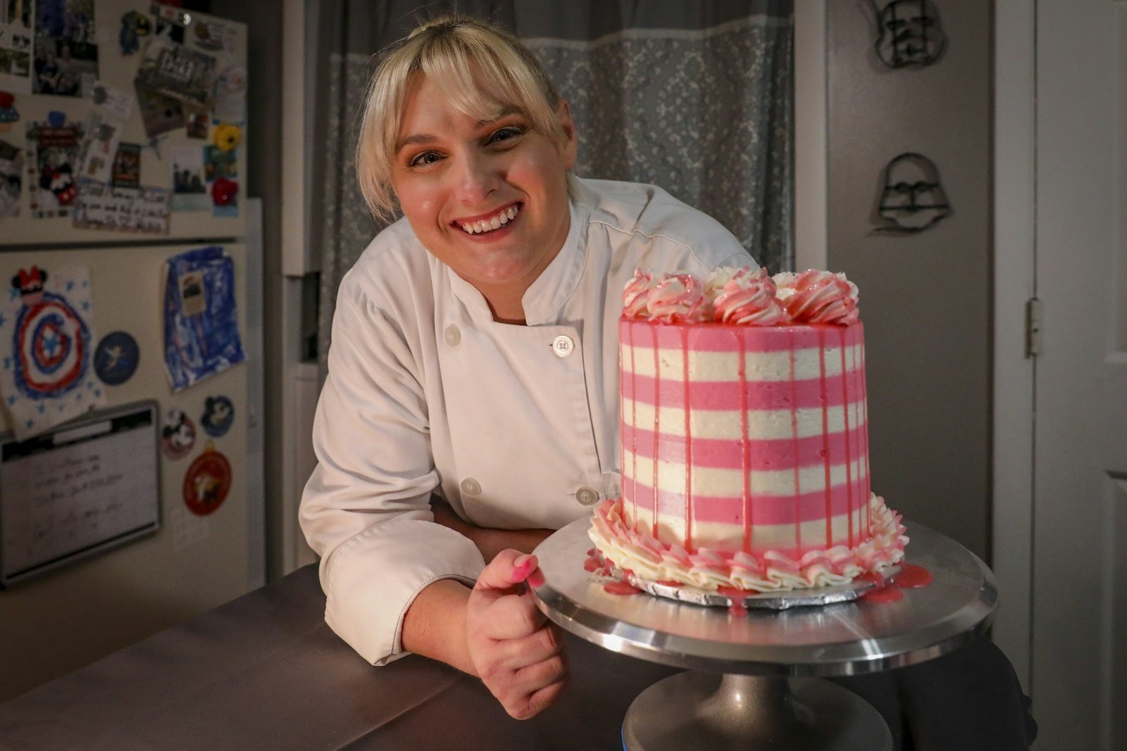 A baker smiling beside a pink striped birthday cake